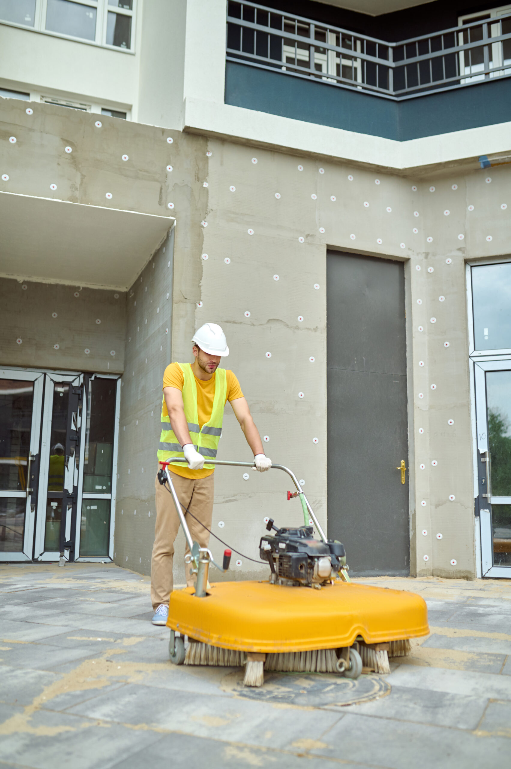 Cleaning. Man in protective helmet and gloves operating special equipment with brush cleaning construction site in new housing complex outdoors